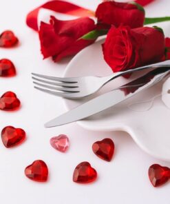 Romantisk bordopdækning med røde roser, bestik og rubinrøde hjerteformede pynte diamanter på hvidt bord.