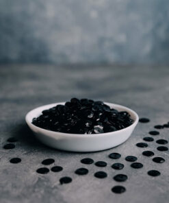 Hvid skål fyldt med sorte palietter, omgivet af spredte palietter på en grå overflade.