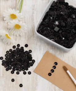 Hvid skål fyldt med sorte palietter, omgivet af spredte palietter på en grå overflade.