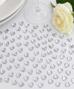 Små sølvhjerter som bordpynt spredt på en hvid dug sammen med tallerken, vinglas og lys rose i et elegant festbord-setup.