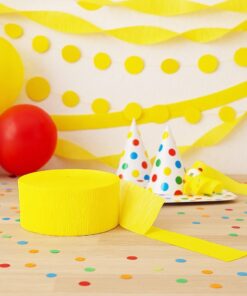 Festlig bordopdækning med gul crepepapirrulle, balloner, guirlander og farvede festhatte i et farverigt partytema.