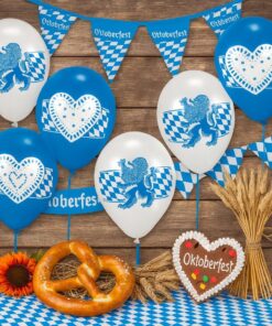 Oktoberfest-stemningsbillede med blå-hvide balloner, bayerske flag, pretzel, honningkagehjerte og hvedeaks foran en trævæg.