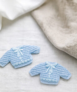 Små lyseblå hæklede trøjer som barnedåbspynt placeret på lyst festbord med tekstiler – dekorativ dåbspynt til baby.