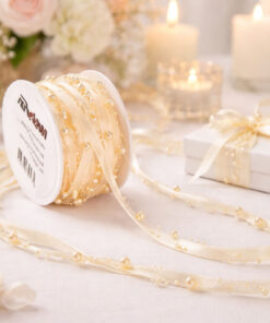 Cremefarvet satinbånd med perler på spole, rullet ud på lyst bord med lys, blomster og gaveæske i elegant festopdækning.