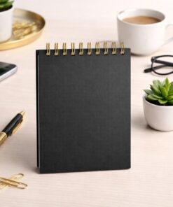 Sort A6 notesbog med guld spiralbinding på skrivebord med pen, kaffe og kontorartikler